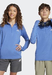 Dvaja jednotlivci nosia modré športové tričká s dlhými rukávmi vyrobené z materiálu, ktorý odvádza vlhkosť, so zipsovým výstrihom a akcentmi loga. Kombinujú ich s šortkami.