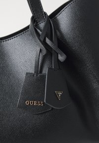 Zwarte leren handtas met textuur, voorzien van twee hangtags; de ene heeft "GUESS" in gouden letters geperst, de andere heeft een klein driehoekig logo.
