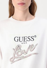 Felpa bianca con logo nero "GUESS®", sfondo triangolare verde e testo "Love" decorato con accenti rosa e argento.