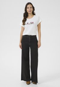 Bela majica s kratkimi rokavi z barvno grafiko čevlja, v kombinaciji z visokim pasom širokimi črnimi hlačami in rjavimi gležnjarji.