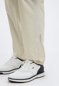 Μπεζ παντελόνια Oakley με ελαστικά μανσέτες φορεμένα με λευκά και μαύρα παπούτσια γκολφ Skechers με κορδόνια σε λευκό φόντο.