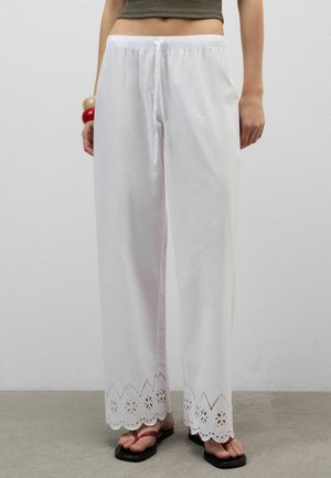 Pantalon blanc à jambes larges avec ourlet festonné à découpe florale porté avec un top court vert foncé et des sandales noires.