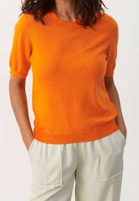 Pullover mit kurzen Ärmeln und Rundhalsausschnitt in leuchtendem Orange, aus weichem Strickstoff, mit einem gerippten Halsausschnitt und Saum, getragen zu hellen Hosen.
