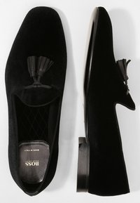 Mocassins en velours noir avec des accents de franges, bout en amande et semelle en caoutchouc noir texturée. L'intérieur doublé présente un motif matelassé.