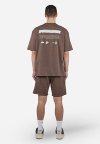 Brun bomull t-skjorte med løs passform, med hvit tekstgrafikk på ryggen. Kombinert med matchende shorts. Brukt med hvite sokker og joggesko.