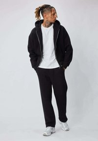 Sweat à capuche noir avec fermeture éclair et poche avant, assorti à un pantalon de jogging coordonné. Un T-shirt blanc est porté en dessous. Des baskets grises complètent le look.