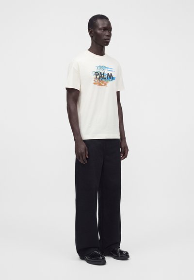 Άνδρας που φοράει λευκό t-shirt με σχέδιο φοίνικα και μαύρο παντελόνι με φαρδιά μπατζάκια, στέκεται μπροστά σε απλό λευκό φόντο.