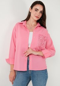 Pink button-up shirt with long sleeves, featuring a lacy pocket accent and fringe detail. Made from a textured fabric with a striped pattern.