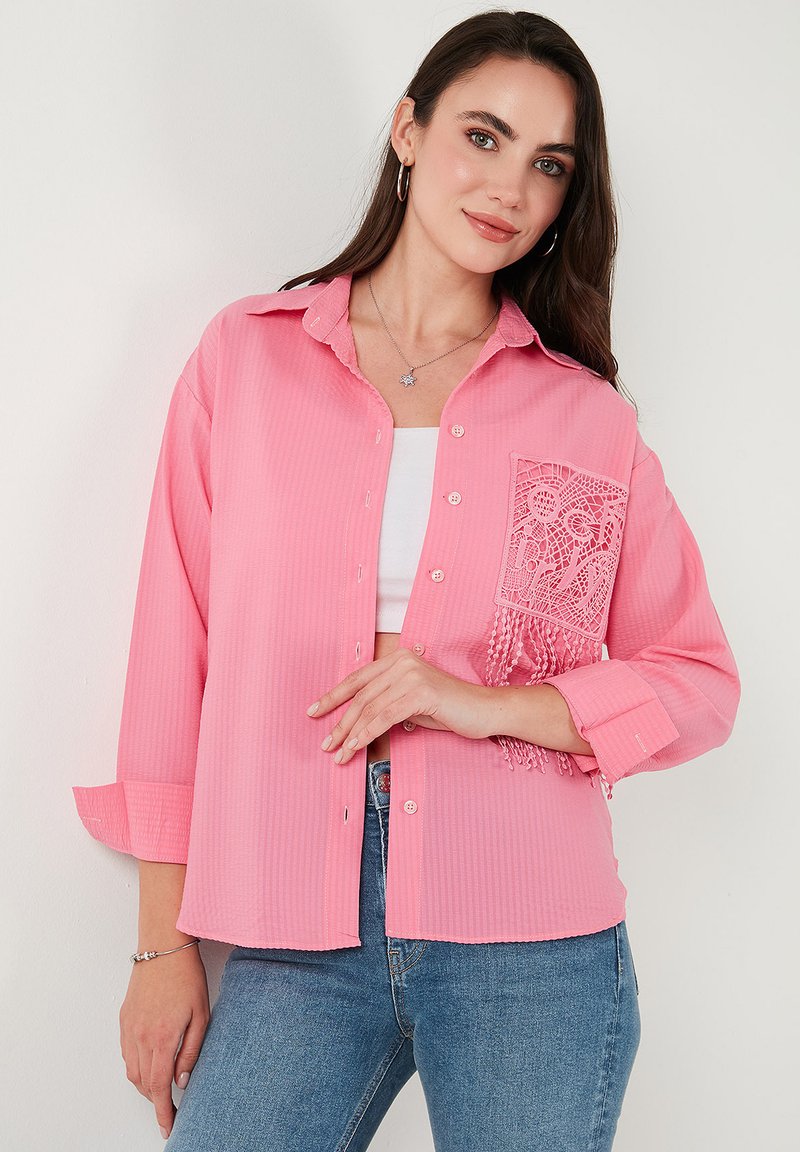 Pink button-up shirt with long sleeves, featuring a lacy pocket accent and fringe detail. Made from a textured fabric with a striped pattern.