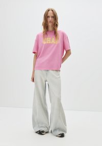 Roze katoenen t-shirt met gele "CHAM" tekst, gecombineerd met lichtblauwe wijde jeans en zwarte sneakers, tegen een witte achtergrond.