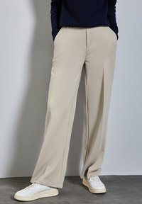 Beige weiteHosen mit glatter Textur, mittiger Bügelfalte und Taschen, kombiniert mit weißen Turnschuhen. Der Hintergrund ist neutral grau.