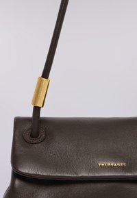 Brązowa skórzana torebka o gładkiej strukturze, z złotym akcentem na pasku i wytłoczonym logo. Kompaktowy prostokątny kształt.