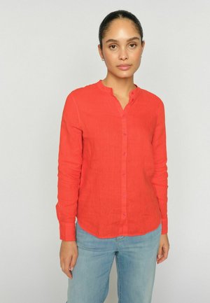 Woman with pulled-back hair wears a red button-up long-sleeve shirt and light blue jeans, standing against a plain light gray background.