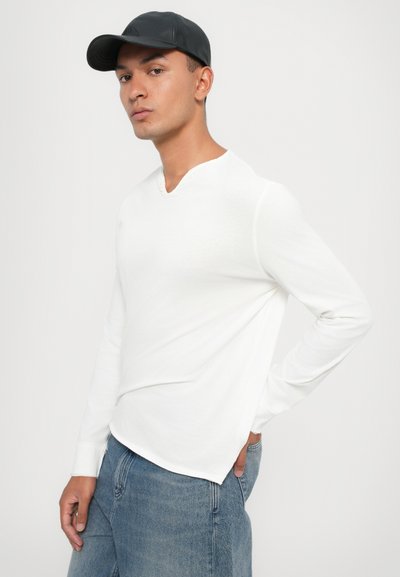 Chemise blanche à manches longues et col en V avec une texture subtile, assortie à un jean en denim bleu. Le modèle porte une casquette noire et se tient devant un fond clair.