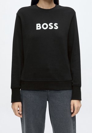 Personne portant un sweat-shirt noir à manches longues avec le mot "BOSS" en lettres blanches sur la poitrine et un jean gris foncé.