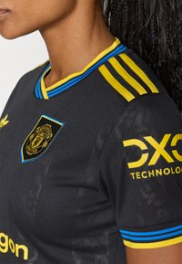 Črna športna majica z rumenimi in modrimi detajli, ki ima v-izrez, tri rumene črte na rami in logo na prsih.