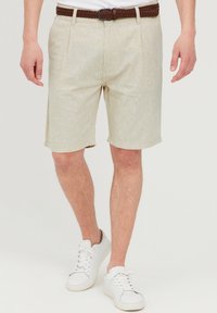 Beige linnedragt shorts med en afslappet pasform, der har en tekstureret overflade og lommer, parret med et brunt vævet bælte og hvide sneakers.