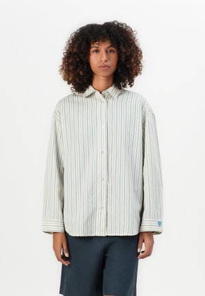 Vrouw met krullend haar draagt een losse, lange mouwen crème shirt met dunne verticale blauwe strepen en donkere shorts, staande tegen een witte achtergrond.