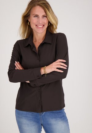 Lachende vrouw met blond haar die haar armen gekruist houdt, draagt een donker shirt met lange mouwen, gouden oorringen, een gouden armband en blauwe spijkerbroek.
