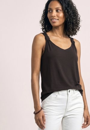 Femme aux cheveux bouclés portant un haut noir sans manches avec des bretelles nouées et un pantalon blanc, regardant légèrement sur le côté et souriant.