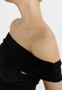 Czarny top typu off-the-shoulder z gładkiego materiału, z miękkim drapowaniem wokół dekoltu i dopasowanym fasonem.