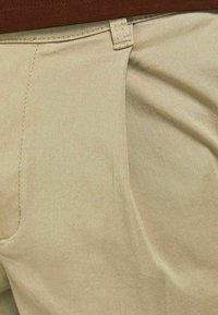 Tan tyg med en slät textur, med en framficka och sydda detaljer. En del av en beige outfit med en brun midjeband synlig ovanför.