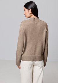 Beige Strickpullover mit hohem Kragen, gerippten Bündchen und Saum, kombiniert mit hellen Hosen, die eine lockere Passform und glatte Textur zeigen.