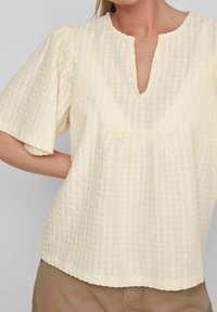 Ljust gult bomullstopp med ett texturerat rutmönster, korta puffärmar och V-ringning. Har sömmar längs kanterna.