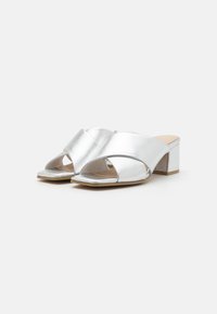 Mules argentés à talons avec une tige en synthétique, dotés d'un design à lanières croisées, d'un bout ouvert et d'un talon carré. Texture lisse.