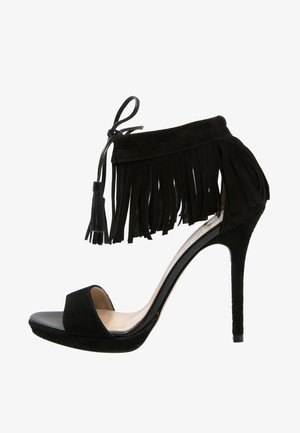 Sandalia de tacón alto negra con parte superior de ante suave, detalle de flecos en el tobillo, diseño de punta abierta y un tacón brillante. Presenta un cierre de lazo.