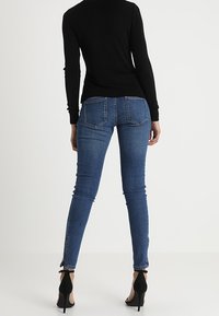 Femme portant un jean bleu ajusté et un haut noir à manches longues, se tenant de dos, avec des chaussures à talons hauts noires sur fond blanc.