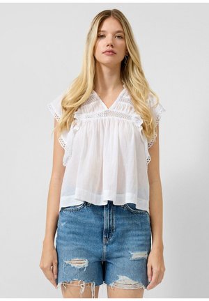 Junge Frau mit langen blonden Haaren trägt eine weiße, mit Spitze verzierte Bluse und ausgewaschene blaue Jeansshorts vor einem schlichten grauen Hintergrund.