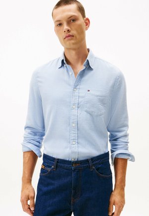 Lys blå skjorte med knapper og lomme, lavet af tekstureret stof, parret med mørkeblå denim jeans. Skjorten har optrukkede ærmer og subtile detaljer.
