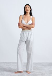 Bela bralette in svetlo sivi prugasti pajama hlače z elastičnim pasom. Model stoji boso, z rokami prekriženimi pred enobarvno ozadje.