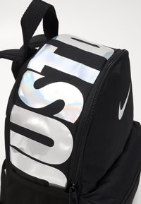 Mochila negra con un logotipo plateado brillante "JUST DO IT". Presenta una tela texturizada, cierre de cremallera y una pequeña pestaña.