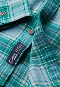 Camicia a quadri verde e blu con accenti azzurri, chiusure a bottoni e un'etichetta in tessuto con i dettagli del marchio, realizzata in morbido cotone.