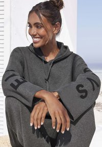 Dunkelgrauer Hoodie mit weiten Ärmeln, der schwarze Schriftzüge auf den Ärmeln trägt. Weicher Stoff, verstellbare Zugkordel und lockere Passform.