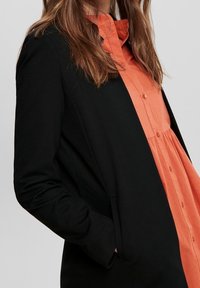 Svart kavaj med långa ärmar, med en slät textur och öppen framkant. Ovanpå en orange skjorta med krage, knappar och lös passform.