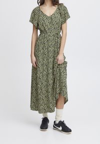 Maxiklänning i mörkgrön med små gula blommönster, korta fjäderlutande ärmar, V-ringning, snäva midja och asymmetrisk nederkant. Bärs med sneakers.