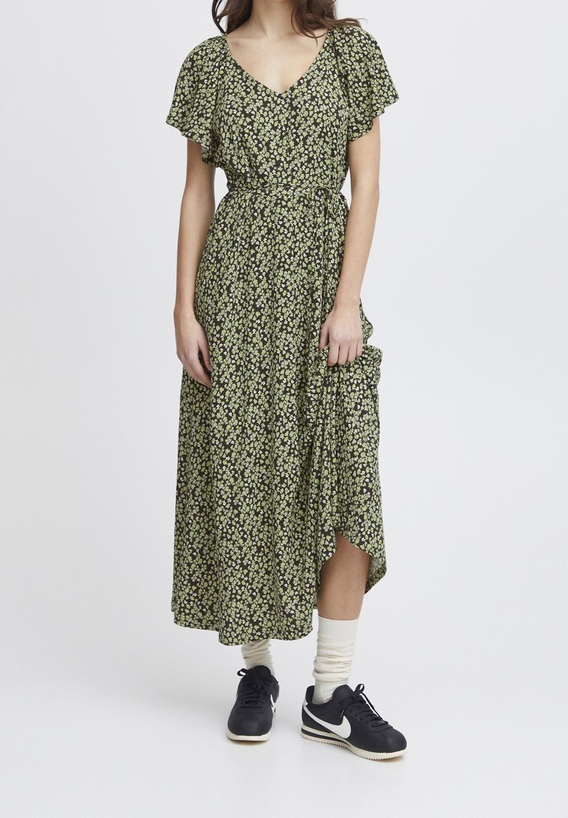 Maxiklänning i mörkgrön med små gula blommönster, korta fjäderlutande ärmar, V-ringning, snäva midja och asymmetrisk nederkant. Bärs med sneakers.