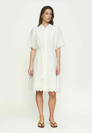 Naine valges aukudega midi pikkuses kleidis, millel on lainelised varrukad ja alumine ääris, vöö seotud vöökohal ning pruunid madalad sandaalid, seistes ühevärvilise tausta ees.