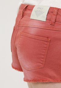 Short en denim rouge avec des ourlets effilochés, une poche arrière et un patch en cuir blanc marqué "ONLY" à la taille.