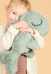 Jong kind dat glimlacht en een zachte, groene pluche walvis knuffelt met gesloten, geborduurde ogen tegen een beige achtergrond.