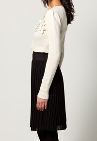 Virkat stil i elfenbensfärgad tröja med ribbade ärmar, parat med en svart veckad kjol. Har strukturerad tyg och lager av volangdetaljer vid bröstet.