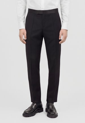 Man die zwarte formele broek, zwarte leren loafers en een wit overhemd met knopen draagt, staand op een effen achtergrond.