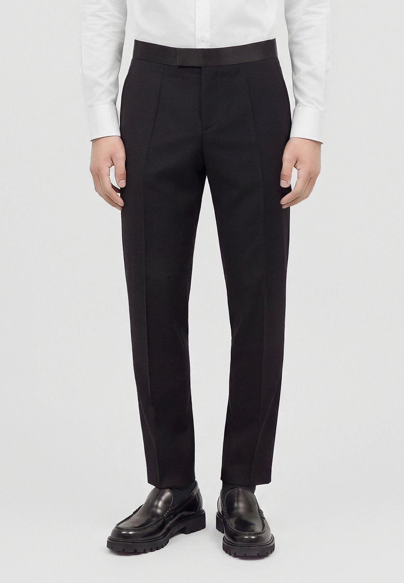 Homme portant un pantalon noir formel, des mocassins en cuir noir et une chemise blanche boutonnée, debout sur un fond uni.