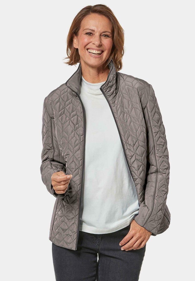 Grijze getailleerde jas met een opstaande kraag en rits. Heeft een gestructureerd patroon en zijzakken. Draagt over een licht shirt.