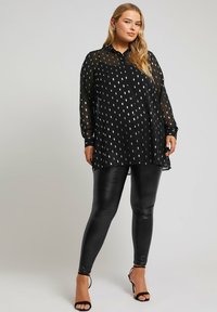Zwarte doorschijnende blouse met zilveren bladeren patroon, lange mouwen, gecombineerd met glanzende zwarte leggings en gespen met hoge hakken.