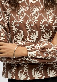 Bruine stof top met ingewikkelde witte bloemenborduursels, opgeblazen mouwen en gerimpelde manchetten. Zilveren armband om de pols. Gelaagde oppervlaktedetails.