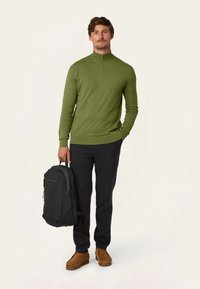 Groene zip-nek trui, zwarte broek, bruine moccasin-stijl schoenen en een zwarte rugzak in één hand. Eenvoudig, modern ontwerp.